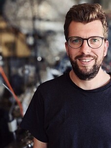 Maximilian Uenzelmann mit dunklem Shirt, Brille und Bart, industrieller Hintergrund.